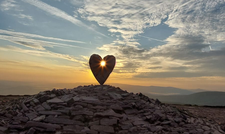 A heart-shaped sculpture on a mountaintop at sunset, with the sun shining through the heart.