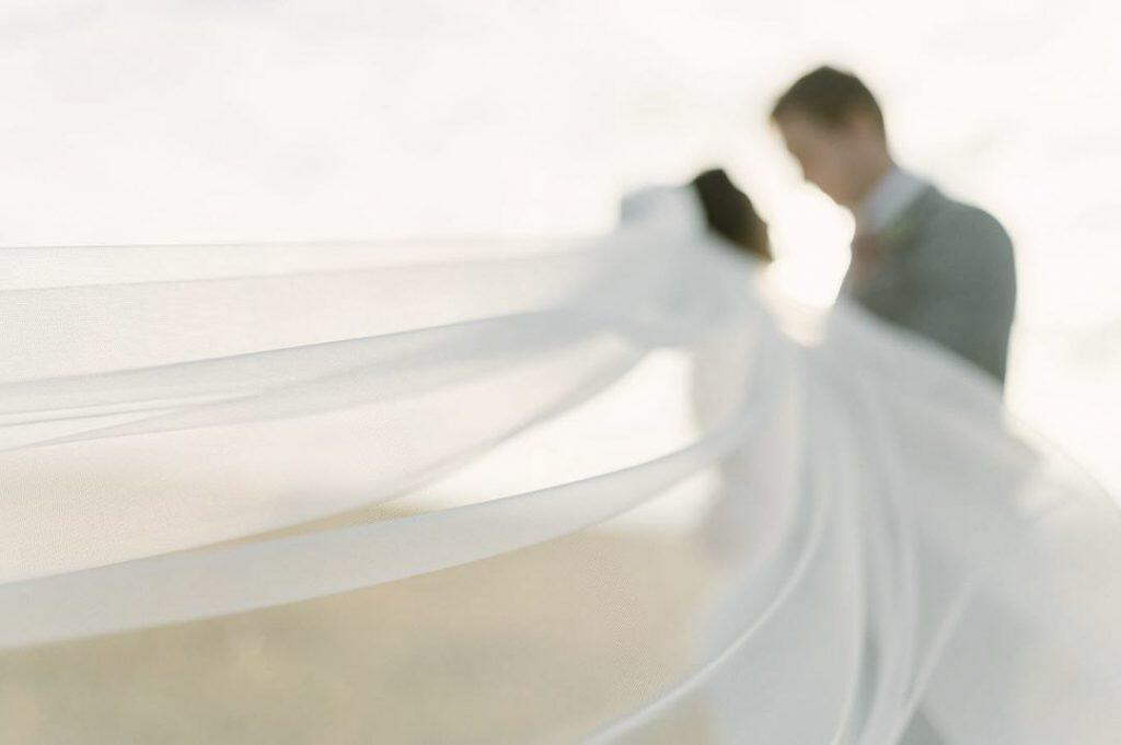 Blurred image of a bride and groom with a flowing wedding veil in the foreground, creating a soft and dreamy effect