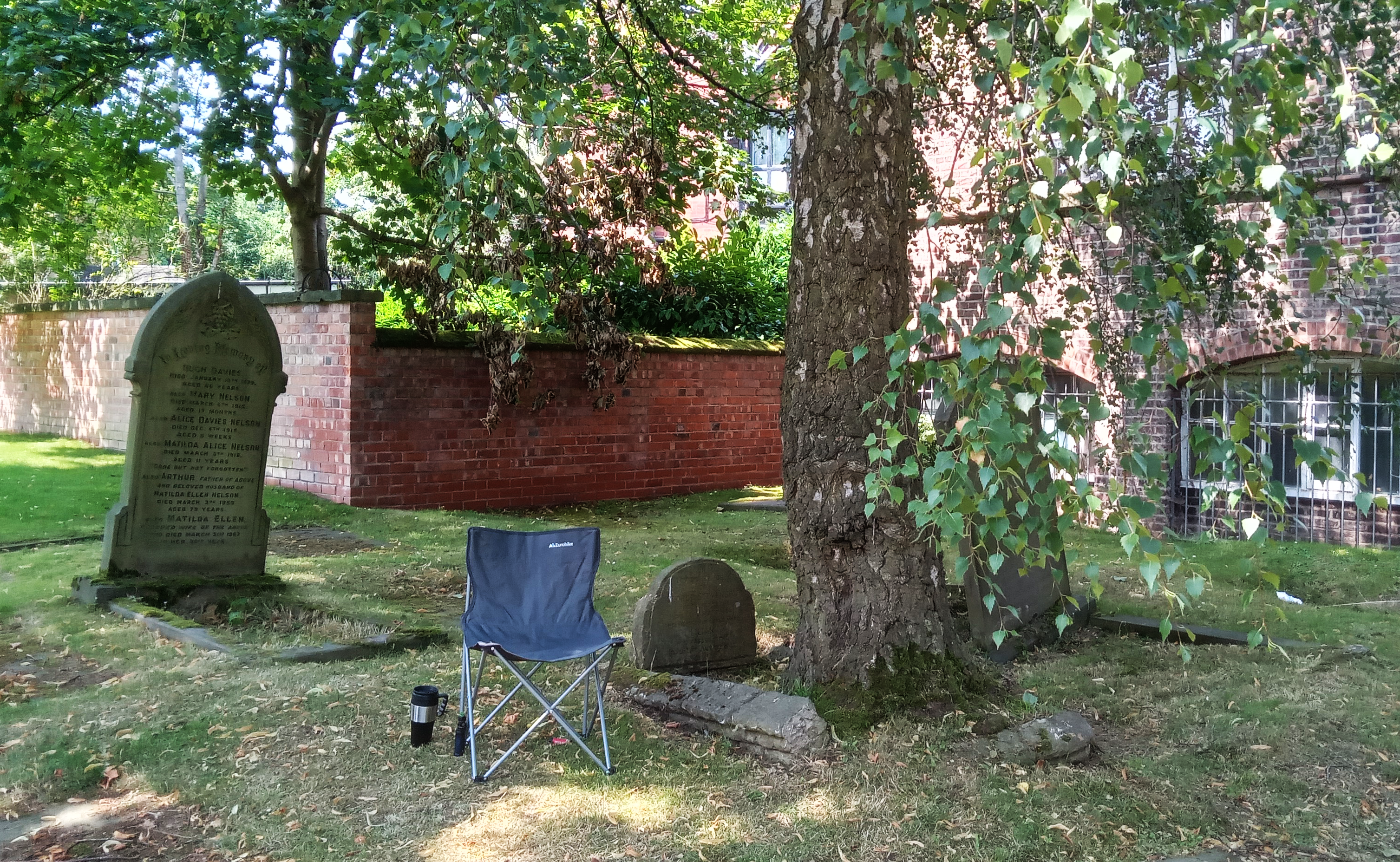 A folding chair sits beneath a large tree in a churchyard, with tombstones and a brick wall in the background.